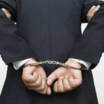 Person handcuffed in suit, escorted by two others.