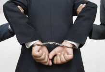 Person handcuffed in suit, escorted by two others.