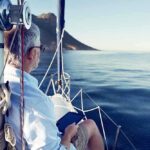 Killer Whales SINK Three Boats in One Week A man sitting on a sailboat, looking out at the calm ocean with mountains in the background