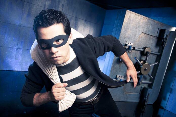 shutterstock_75825736.jpg A masked thief in a striped shirt holding a bag while emerging from a bank vault