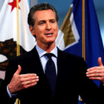 Newsom’s Bonehead IDEA to Cover HIS Fiscal Mismanagement Man speaking with flags in the background