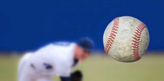 A baseball in mid-air with a pitcher in the background preparing to throw