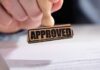 A hand holding a wooden stamp with the word APPROVED above a document
