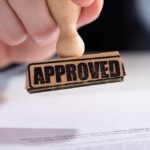 A hand holding a wooden stamp with the word APPROVED above a document