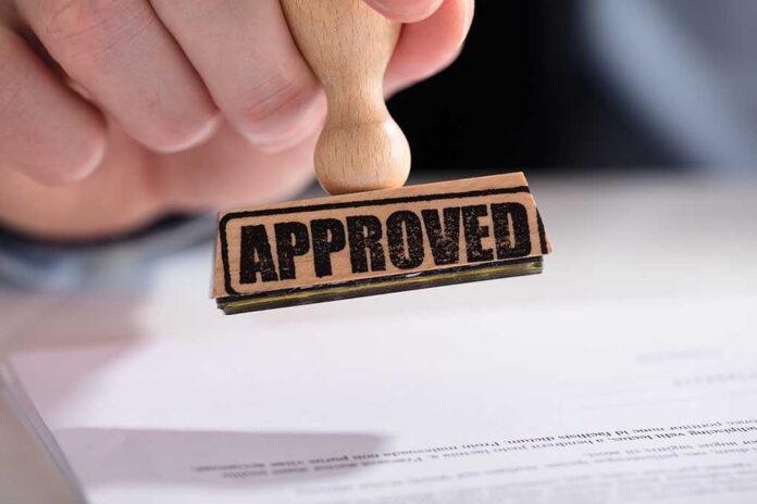 A hand holding a wooden stamp with the word APPROVED above a document