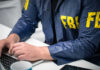 Person in FBI jacket typing on a laptop.