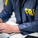Manifesto LEAKED — FBI Reveals Assassination Hit List Person in FBI jacket typing on a laptop.