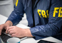 Person in FBI jacket typing on a laptop.