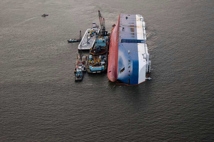 1540854386 Capsized ship with cranes and tugboats around it.