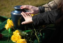 Hands touching a blue cremation urn beside yellow roses