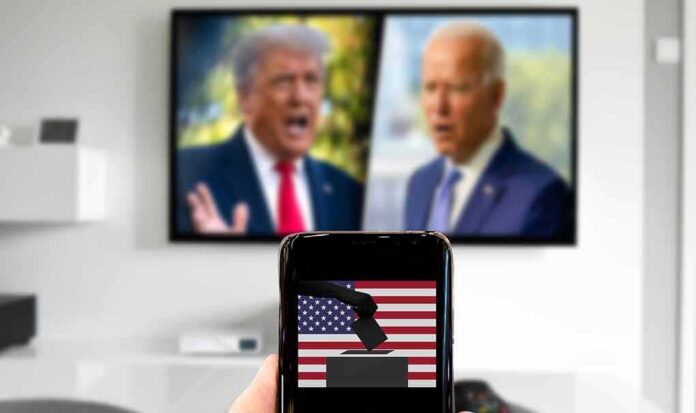 shutterstock_1823317808 Smartphone with ballot icon held before a TV showing two blurred politicians