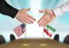 Two hands reaching to shake above US and Iran flags on a conference table